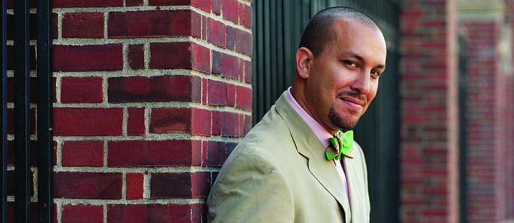 Josef Sorett is an associate professor of religion and African American Studies and director of Columbia’s Center on African American Religion, Sexual Politics, and Social Justice. Photo by Gabriel Cooney Daylight: Columbia University Religion Professor Josef Sorett leans against a red brick pillar, smiling slightly and wearing a pale green suit, pink button-down shirt, and bright green bow tie; photo by Gabriel Cooney.