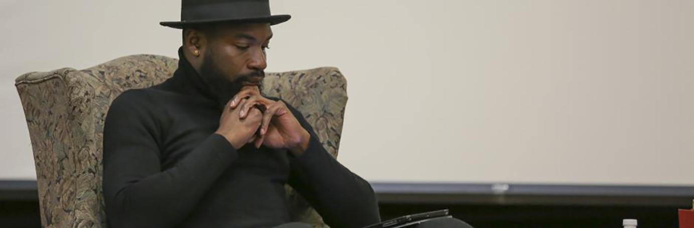 A brown-skinned man wearing a black turtle-neck and stylish hat sits contemplatively in an armchair with his finger interlaced